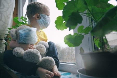 Un enfant portant un masque ayant une peluche dans la main regarde par la fenetre